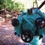 Turquoise engine block with three black pulley wheels mounted on front, displayed outdoors with vegetation and stone wall background