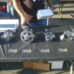 Four polished chrome automotive alternators and starters displayed on a black table with small identification plaques