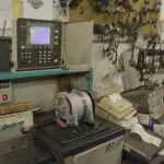 Industrial lathe with CNC control panel and motor mounted on turquoise workbench in electronics repair workshop with pegboard tool storage.