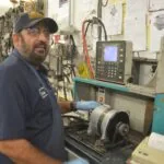 Technician in navy work shirt and cap standing at CNC lathe machine with control panel and shop drawings on wall behind
