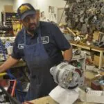 Technician in blue apron and cap working on an alternator in a cluttered auto shop with engine parts and tools visible