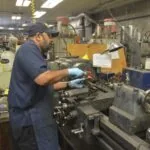 Mechanic in blue shirt and cap working on engine components at workbench in auto shop