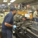 Worker in blue shirt and cap operating a lathe machine in an industrial workshop with equipment and tools visible