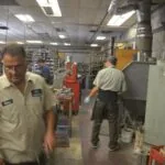 Two workers in uniform shirts inside an auto repair shop with tools, equipment, and shelving