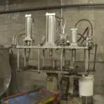 Industrial distillation or filtration apparatus with stainless steel cylinders, piping, and gauges mounted on wall with concrete blocks