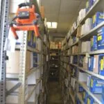 Orange chainsaw hanging on left shelf in narrow warehouse aisle lined with boxed automotive parts and supplies
