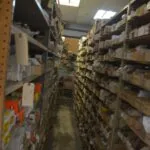 Long narrow warehouse aisle lined with cardboard boxes stacked on metal shelving on both sides, fluorescent ceiling lights overhead