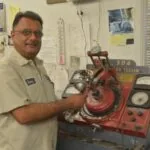 Man in light colored work shirt standing next to a red automotive diagnostic testing machine in a shop