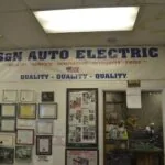 Interior of S&N Auto Electric shop with signage emphasizing quality, service, innovation, integrity, and pride on white wall with framed certificates and photos below