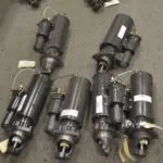 Six black electric starter motors arranged on gray floor, each labeled with white tags